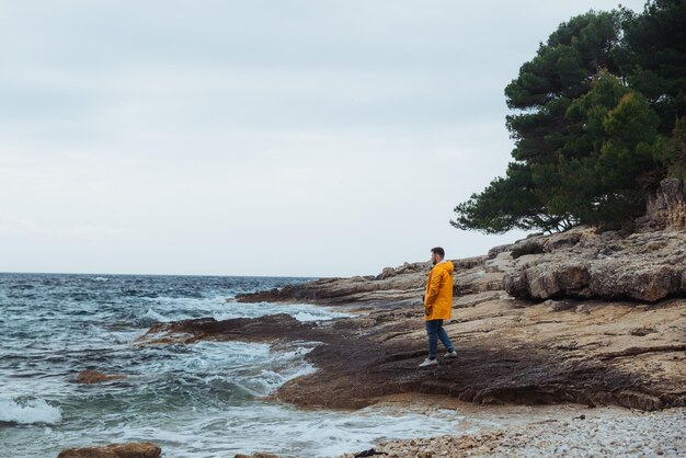 Mann, der am felsigen Strand bei bewölktem, windigem Wetter mit gelbem Regenmantel spazieren geht