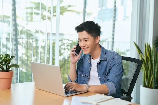 Mann, der am Handy spricht und von zu Hause aus arbeitet. Porträt eines nachdenklichen asiatischen Geschäftsmannes mit Laptop-Computer, Planungsprojekt, Kommunikation im modernen Büro