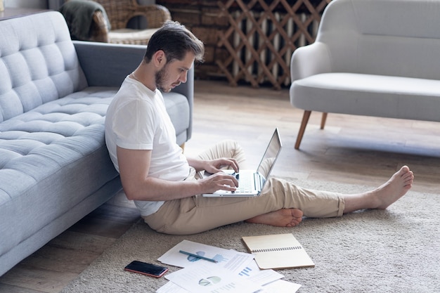 Mann, der am Laptop arbeitet, der auf dem Boden mit seinem Tagebuch und offiziellen Papieren zu Hause sitzt