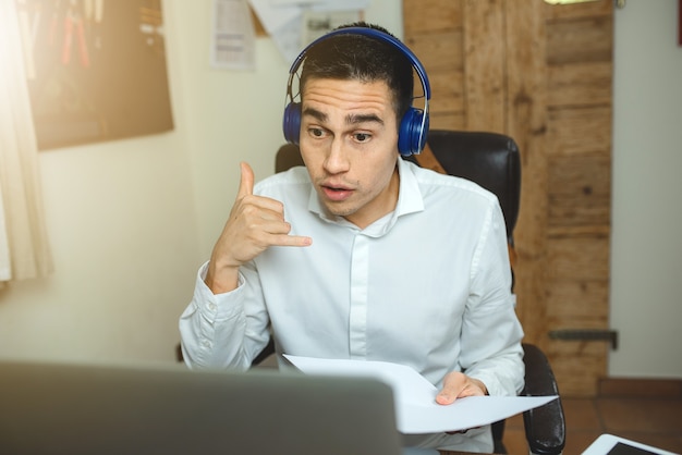 Mann, der am Laptop arbeitet, einen Videoanruf mit Kopfhörern hat und Papiere hält. Mit der Hand diskutieren und Gesten machen.