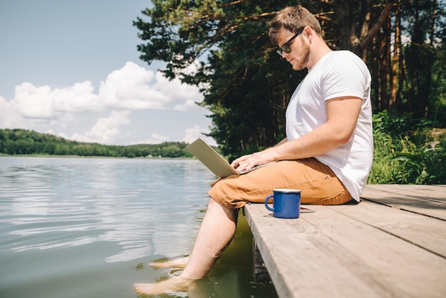 Mann, der am Laptop arbeitet, während er auf hölzernen Stegbeinen im Flusswasser sitzt