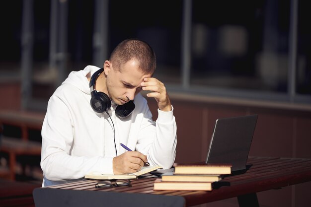 Mann, der an einem laptop arbeitet und in ein notizbuch schreibt, das auf der straße an einem tisch sitzt. soziale distanzierung während des coronavirus