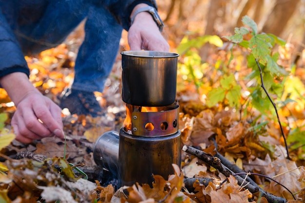 Mann, der an einem offenen Feuer im Wald kocht Ein Tourist erhitzt Wasser in einem Metallbecher auf einem Feuer in einem Herbstwald