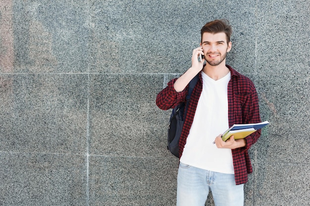 Mann, der auf dem Smartphone auf grauem Hintergrund im Freien spricht und während der Pause Notizbücher in der Hand hält, sich auf dem Universitätscampus ausruht. Bildungs- und Kommunikationskonzept, Kopienraum