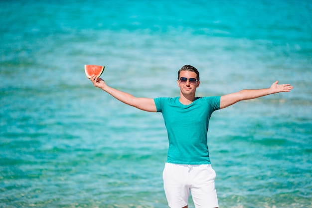 Mann, der auf dem Strand an einem Sommertag lächelt, der mit einer Scheibe Wassermelone in der Hand lächelt