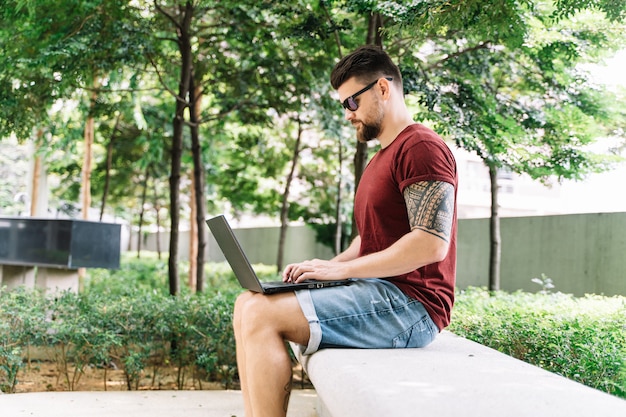 Mann, der auf einer Bank mitten in einem Park sitzt, der entfernt mit seinem Laptop arbeitet