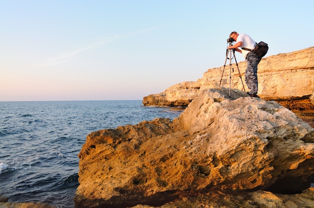Mann, der auf Felsen steht und Foto mit Stativ über Schwarzem Meer macht