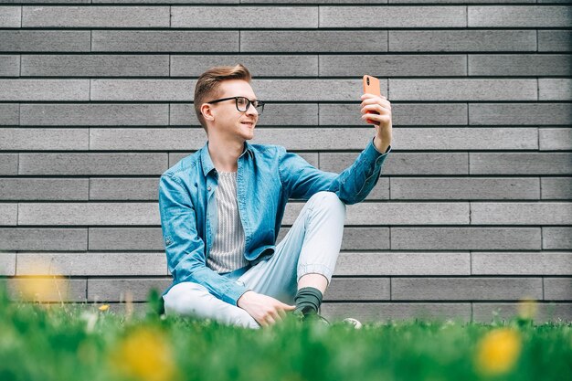 Mann, der auf grünem Gras sitzt und Smartphone auf einem grauen Wandhintergrund betrachtet