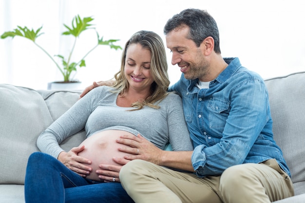 Mann, der auf Sofa sitzt und den Magen der schwangeren Frau hält