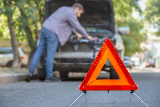 Mann, der Auto auf der Straße repariert. Autopanne während der Fahrt. Fahrer schaut auf Panne unter der Motorhaube des Autos. Rotes dreieckiges Stoppschild auf der Straße.