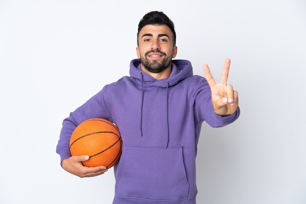 Mann, der Basketball über isolierte weiße Wand spielt, die lächelt und Siegeszeichen zeigt