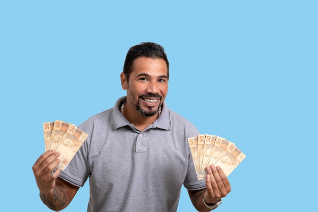Mann, der brasilianisches Geld lächelnd hält, betrachtet Kamera-Werbekonzept isoliert auf blauem Hintergrund