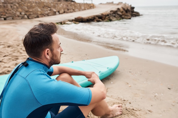 Mann, der das Meer mit Paddelbrett auf dem Sand betrachtet