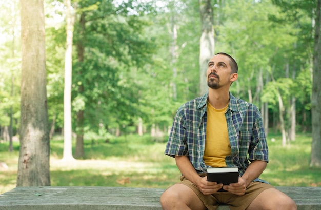 Mann, der ein Buch liest und hält und in einem grünen Park im Freien aufschaut. Studentisches Denken, Religion.