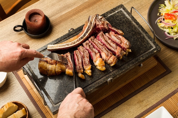 Mann, der ein gegrilltes Steak mit Knochen auf einem Kochstein in einer Draufsicht schneidet
