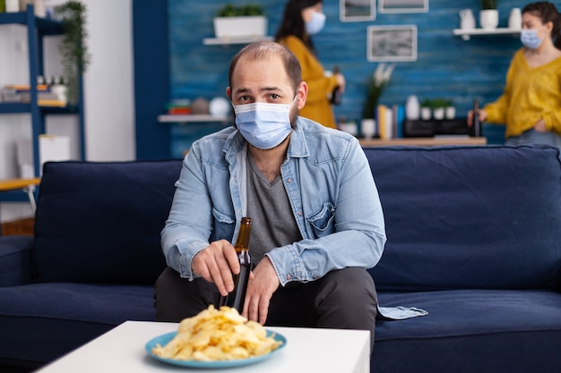 Mann, der eine Bierflasche hält, die sich auf der Couch entspannt, verbringt Zeit mit Freunden, die soziale Distanz mit Gesichtsmaske im Wohnzimmer halten und nachdenklich in die Kamera schauen, um die Ausbreitung des Coronavirus zu verhindern.