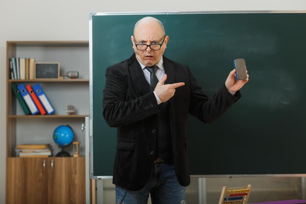 Mann, der eine Brille trägt und neben der Tafel im Klassenzimmer steht und den Unterricht erklärt, der einen Schwamm hält, der mit dem Zeigefinger darauf zeigt und unzufrieden aussieht