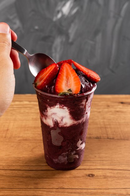 Mann, der einen Löffel brasilianischen Frozen Açai Berry Ice Cream Smoothie in Plastikbecher mit Erdbeeren und Kondensmilch nimmt. Auf Sommerhintergrund. Frontansicht für Speisekarte und Social Media