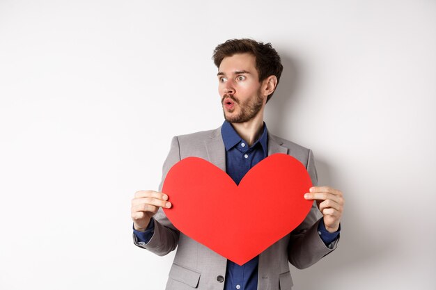 Mann, der erstaunt auf jemanden schaut, der schön ist, großen roten Herzausschnitt hält und am Valentinstag über weißem Hintergrund steht. Speicherplatz kopieren