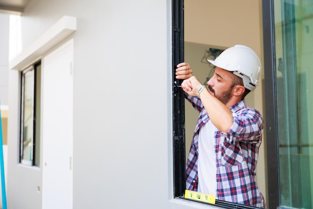 Mann, der Fenster in einer neuen Hausbaustelle installiert. Hispanischer Bauarbeiter mit Schutzhandschuhen und Hardhat-Helm