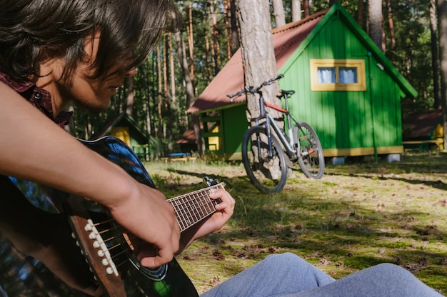 Mann, der Gitarre spielt. Vor dem Hintergrund eines Waldhauses in der Natur.