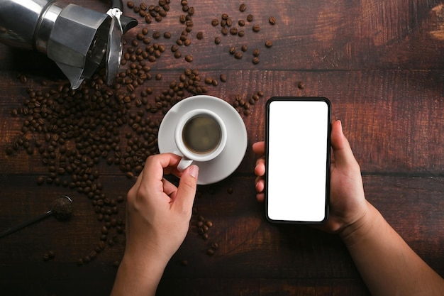 Mann, der Handy und Kaffeetasse auf hölzernem Hintergrund mit gerösteten Kaffeebohnen hält