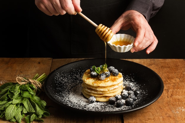 Mann, der Honig auf einen Haufen Pfannkuchen mit Blaubeeren auf einem schwarzen Teller eintaucht