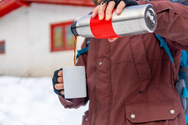 Mann, der im Winter im Schnee neben einer Hütte Kaffee aus einer heißen Thermoskanne trinkt