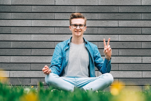 Mann, der in der Pose der Meditation auf grünem Gras auf dem Hintergrund einer grauen Wand sitzt