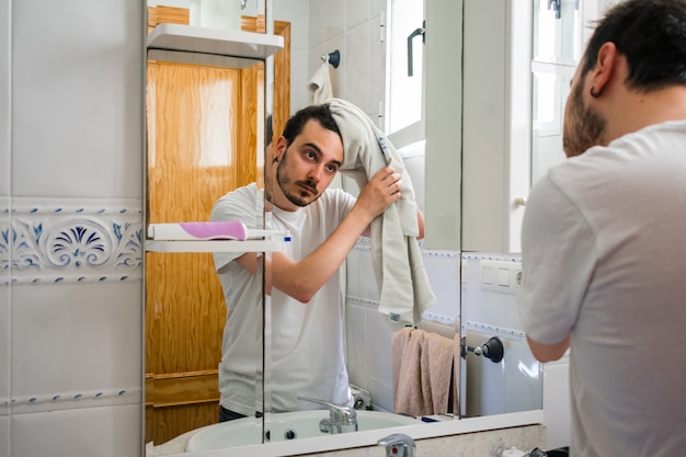 Mann, der in einem Spiegel im Badezimmer sich betrachtet. Er trocknet sich die Haare mit einem Handtuch.