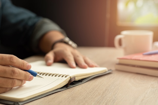 Mann, der in Notizbuch mit Stift und Kaffeetasse auf Holztisch schreibt