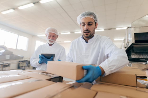 Mann, der Kisten stapelt, während anderer Mann steht und Tablette verwendet. Innenausstattung der Lebensmittelfabrik.