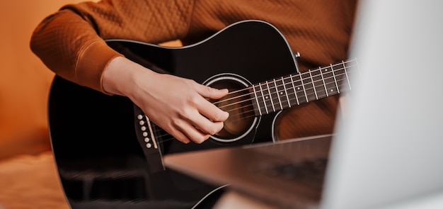 Mann, der lernt, Gitarre mit Hilfe des Online-Lernens zu Hause zu spielen. Kerl sitzt am Tisch mit Laptop und schwarzer Gitarre