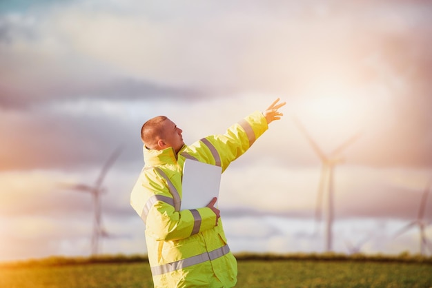 Mann, der mit einem Tablet auf einer Windmühlenfarm arbeitet, die Strom erzeugt, saubere Energie Mann mit einem Laptop mit Windturbinenhintergrund