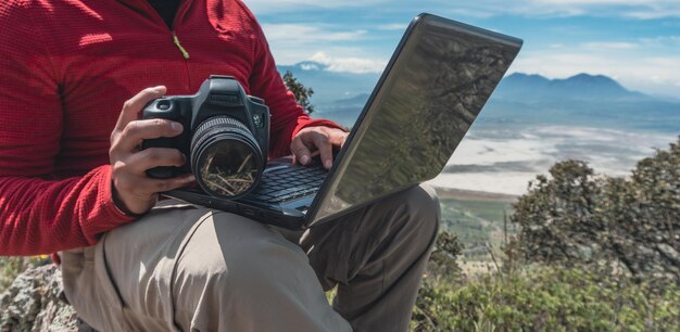 Mann, der mit Laptop und Kamera auf dem Gipfel des Berges arbeitet