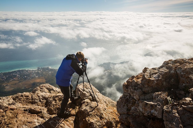 Mann, der mit Stativ und Kamera auf einem hohen Berggipfel über Wolken, Stadt und Meer steht