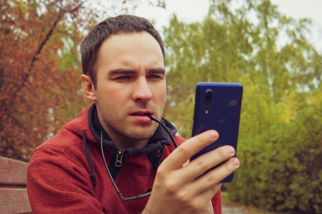 Mann, der neugierig ein Smartphone hält, das draußen im Garten sitzt Ein junger Mann schaut empört auf den Bildschirm seines Smartphones Lesen Sie schlechte Nachrichten, um Geschäftsprobleme online zu lösen