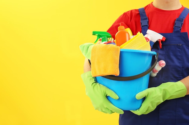 Mann, der Plastikeimer mit Bürsten, Handschuhen und Reinigungsmitteln auf gelbem Hintergrund hält