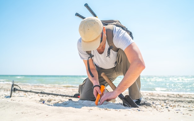 Mann, der Sand gräbt, wenn an sonnigem Tag etwas mit Metalldetektor an der Seeküste gefunden wird.