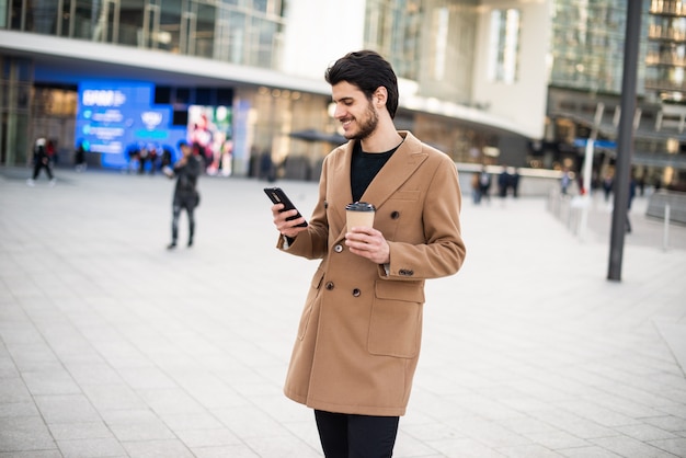 Mann, der sein Handy benutzt und eine Tasse Kaffee hält, während er in einer Stadt geht