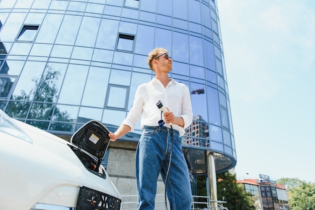 Mann, der sein luxuriöses Elektroauto an der Außenstation vor modernen neuen Stadtgebäuden auflädt