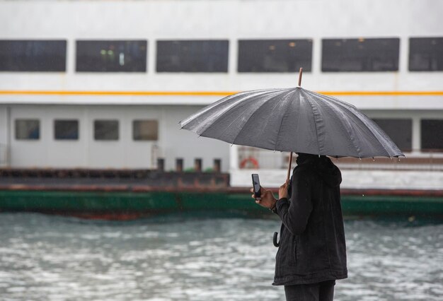 Mann, der sein Telefon am Meer mit schwarzem Regenschirm anschaut