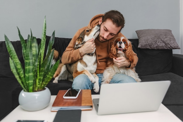 Mann, der sich mit Hunden umarmt, während er am Laptop arbeitet, während er zu Hause auf dem Sofa sitzt