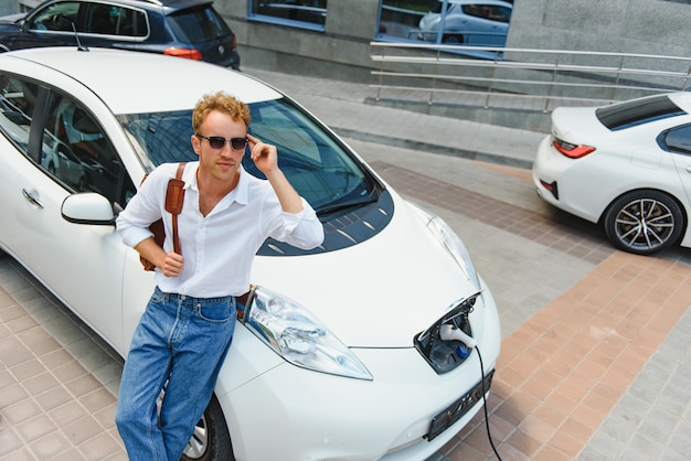 Mann, der Stromladekabel für Elektroauto im Parkplatz im Freien hält. Und er wird das Auto an die Ladestation auf dem Parkplatz in der Nähe des Einkaufszentrums anschließen