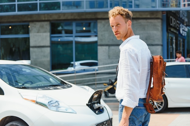 Mann, der Stromladekabel für Elektroauto im Parkplatz im Freien hält. Und er wird das Auto an die Ladestation auf dem Parkplatz in der Nähe des Einkaufszentrums anschließen