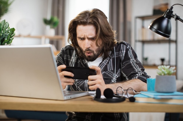 Mann, der während der Pause in einem hellen, modernen Büro oder Zuhause Videospiele auf seinem Smartphone spielt