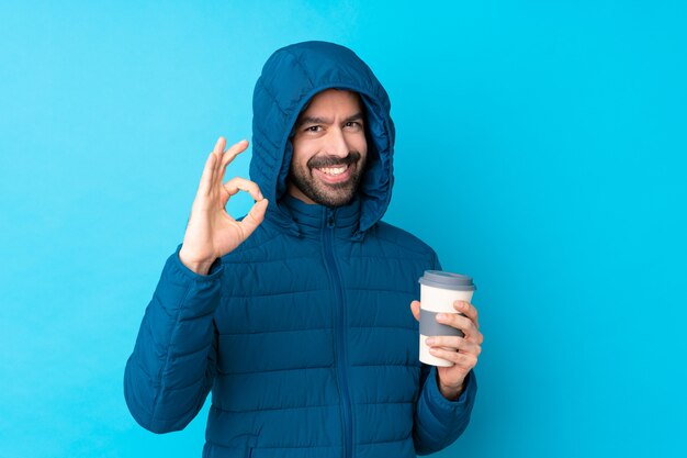 Mann, der Winterjacke trägt und einen Kaffee zum Mitnehmen über isolierte blaue Wand hält, die ok Zeichen mit Fingern zeigt