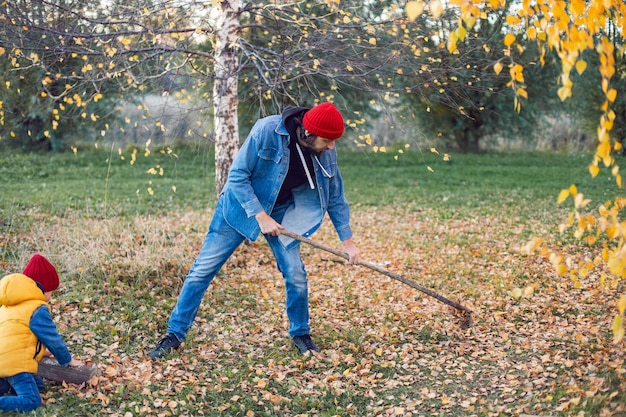 Mann, ein Vater und ein Junge, ein Kind in einer roten Strickmütze säubert Herbstblätter mit Rechen bei Sonnenuntergang hilft im Haus