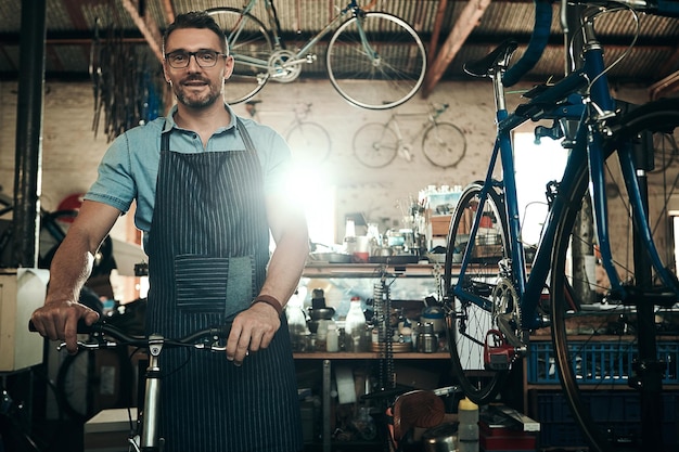 Mann Fahrradladen und Porträt für Wartungsarbeiten Startup mit Transportdienstleistung Fahrradmechaniker und männlicher Unternehmer in der Werkstatt mit Brille Glück für ein nachhaltiges Geschäft mit Schürze