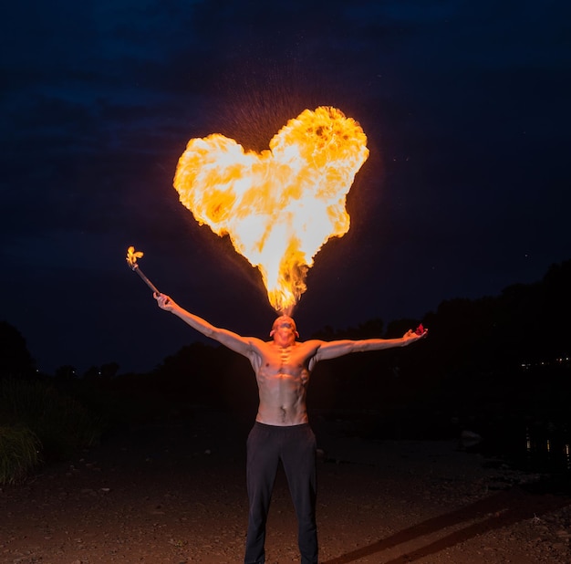 Mann Feuerschlucker bläst nachts eine große Flamme in Herzform aus dem Mund